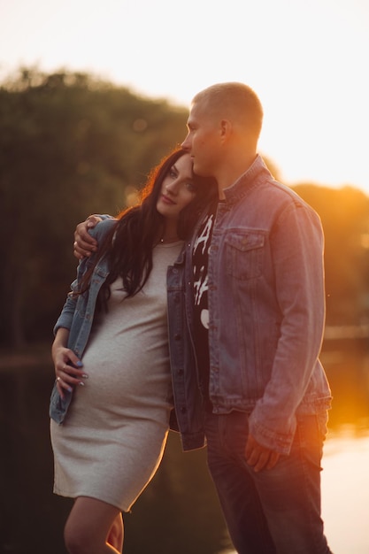 Beautiful pair expecting little child Lovely parents standing near lake hugging each other and posing Pretty pregnant woman touching belly and enjoying motherhood Concept of love and pregnancy