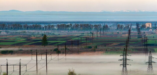 Beautiful panorama of long electric high voltage power line rows stretching to horizon through spring fields and trees under dense morning fog on distant Carpathian mountains range background.