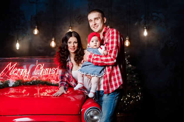 beautiful parents holding their little cute daughter in their arms having fun near retro car in studio. Christmas family look. New Year's scenery.