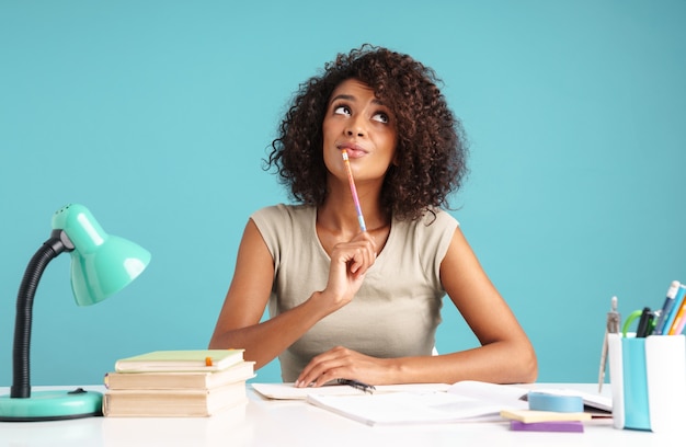 Photo beautiful pensive young african businesswoman casually dressed sitting at the desk isolated over blue wall, studying