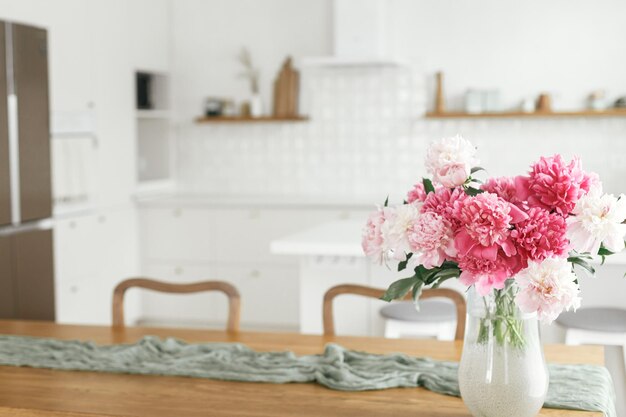 Photo beautiful peonies in vase on wooden table on background of stylish white kitchen with appliances in new scandinavian house modern kitchen interior and summer floral arrangement