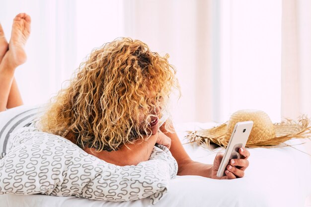 Bella gente bionda capelli lunghi ricci giovane donna usa la tecnologia moderna telefono posa sul letto