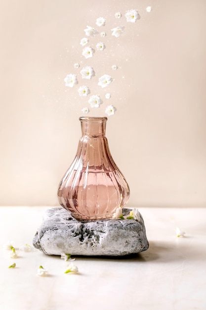 Beautiful pink transparent bottle of perfume standing on textured stone on white marble table Small white flowers Kalanchoe flowing into Perfume making concept with place for design label