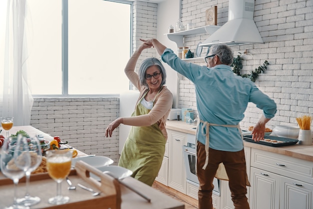 自宅で健康的な夕食を準備しながら踊り、笑顔のエプロンで美しい遊び心のある年配のカップル