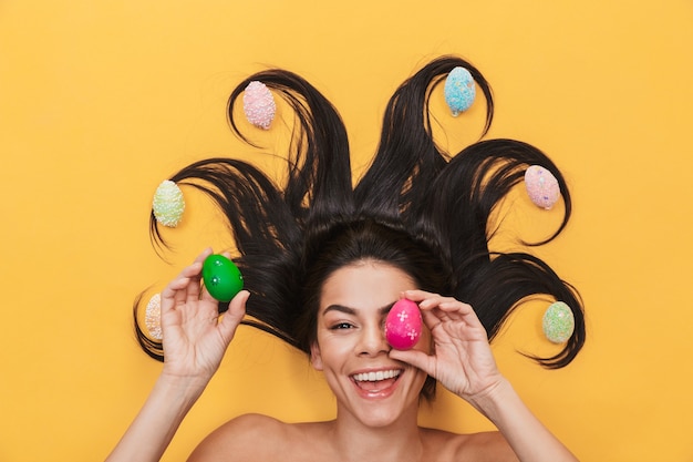 beautiful pleased happy young woman lies isolated on yellow wall over easter eggs covering eyes