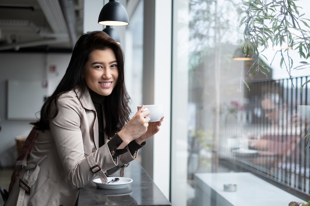 Photo beautiful portrait asian girl sitting on counter bar in coffee shop holding coffee cup with smile looking at camera.