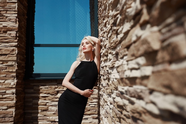 Beautiful portrait of a blonde woman against a brick wall on the street in a black tight dress.