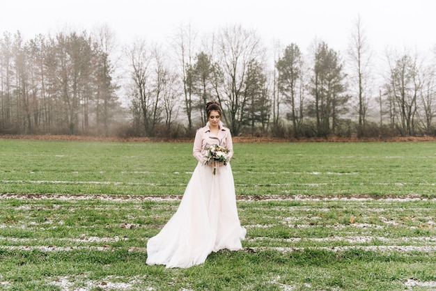 A beautiful portrait of the bride in a stylish dress with a winter bouquet of roses cotton and spruce against a green field