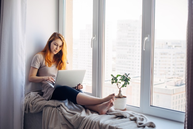 Beautiful positive girl using laptop, sitting on the windowsill in city apartment. Young red-haired woman working at home. Freelance concept.