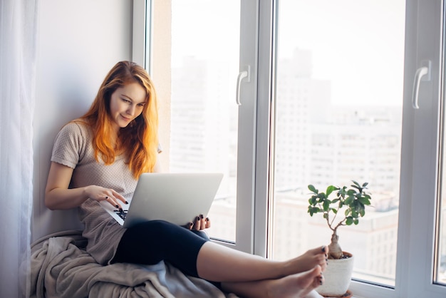 Beautiful positive girl using laptop, sitting on the windowsill in city apartment. Young red-haired woman working at home. Freelance concept.