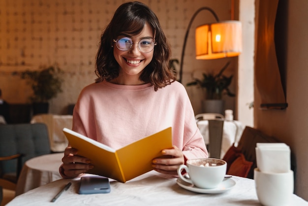 a beautiful positive smiling brunette young woman indoors in cafe reading book.