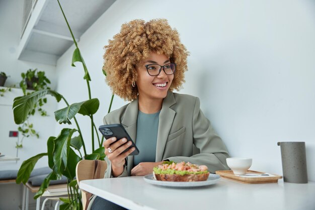Beautiful positive successful businesswoman dressed in formal\
outfit holds mobile phone checks notification on cellphone browses\
internet poses in cozy cafeteria has lunch break during\
workday