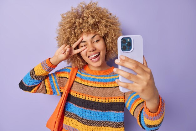 Beautiful positive woman makes peace gesture over eye sticks
out tongue winks eye poses at smartphone camera makes selfie
dressed in knitted jumper carries small bag isolated over purple
wall