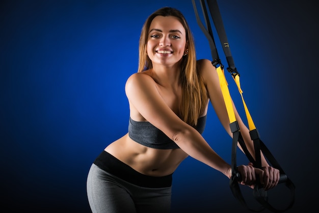 Beautiful positive young girl fitness model posing in studio holding on to hanging straps