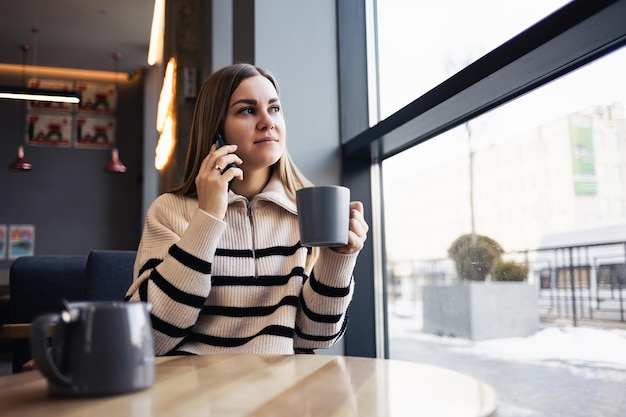 Bella giovane donna positiva in abiti casual che sorride, beve una tazza di caffè e tiene un telefono in mano mentre trascorre del tempo nella caffetteria