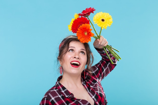 格子縞のシャツと青い表面にポーズをとって美しい明るいガーベラの花を嗅ぐ包帯で美しいポジティブな若い女性