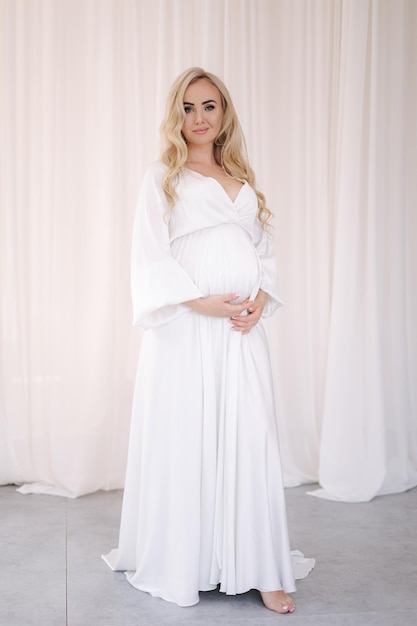 Beautiful pregnant blond hair woman in elegant white dress posing to photographer in studio