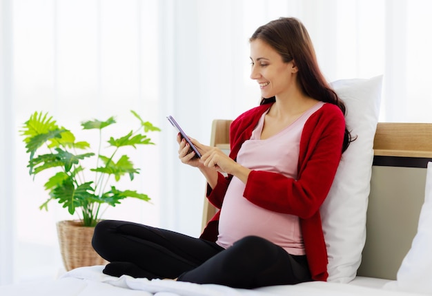 A beautiful pregnant caucasian woman is happily using her smartphone in bed while resting