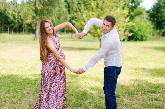 Beautiful pregnant couple happy together expecting a child. Man and woman walking in the park showing heart with hands. Share the love and family, love concept.