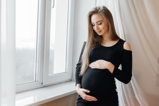 A beautiful pregnant girl is standing in a room near the window