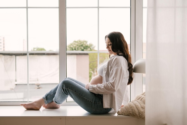 Beautiful pregnant girl with curls in a white shirt and jeans in a bright studio with a stylish interior the concept of a happy pregnancy and family