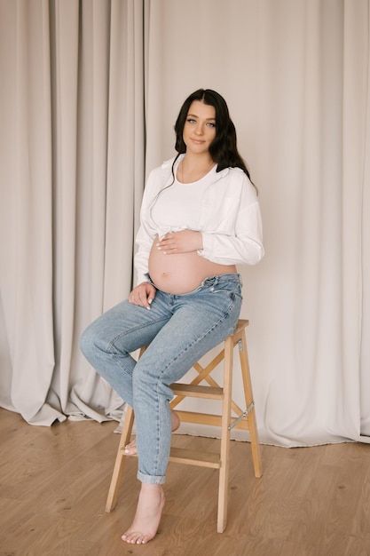 Beautiful pregnant girl with curls in a white shirt and jeans in a bright studio with a stylish interior the concept of a happy pregnancy and family