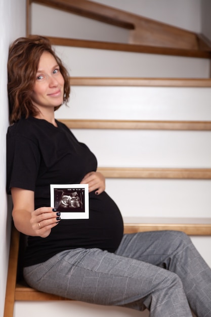 Красивая беременная улыбающаяся кавказская кудрявая женщина сидит на деревянной лестнице и показывает УЗИ