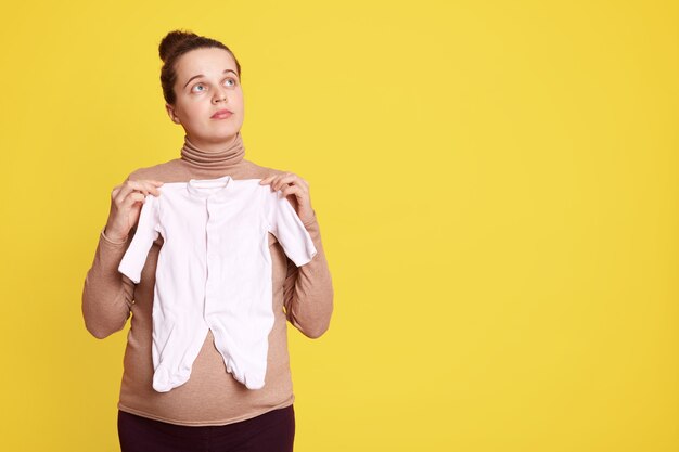 Beautiful pregnant woman in casual clothing, looking away and holding babies clothes in hands, expectant mother with hair bun standing isolated over yellow wall.