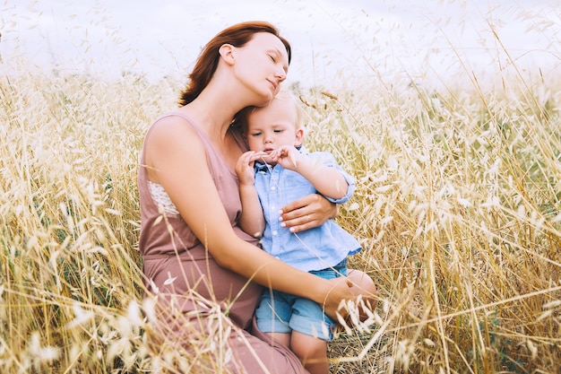 Beautiful pregnant woman and child having fun on wheat field with haystacks at summer day on nature