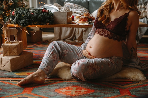 クリスマスツリーの近くの花屋の上に座って快適な服で美しい妊娠中の女性。