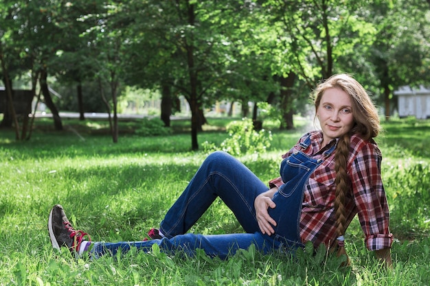 Beautiful pregnant woman in denim overalls sitting on the grass in the park