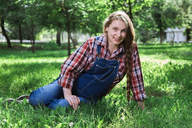 Beautiful pregnant woman in denim overalls sitting on the grass in the park