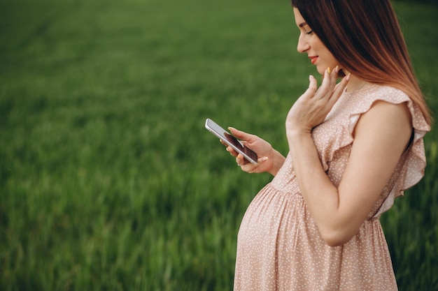 緑の野原に立っている間、美しい妊婦は電話を楽しんでいます