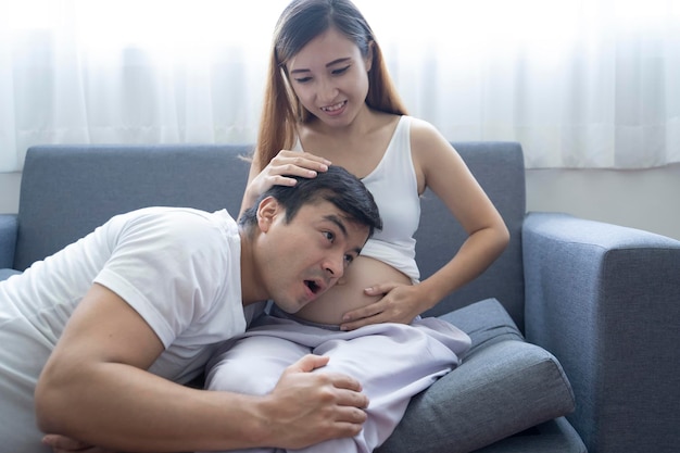 Beautiful pregnant woman and her handsome husband are smiling while spending time together in sofa Man is listening to baby in belly