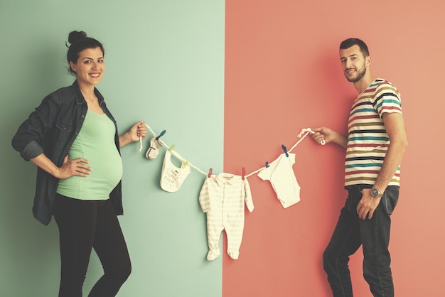 Beautiful pregnant woman and her husband expecting baby holding baby bodysuits and smiling over colorful background