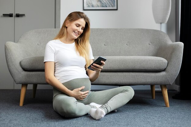 Beautiful pregnant woman holding mobile phone touching belly shopping online sitting at home