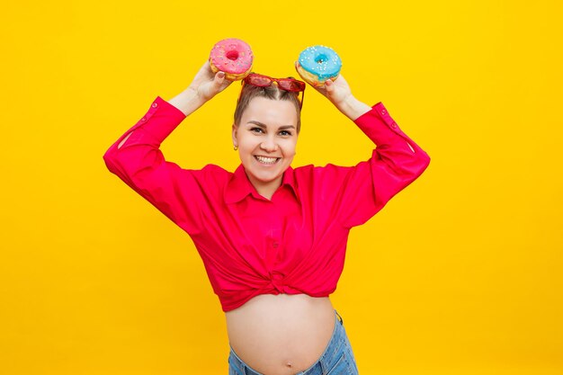 A beautiful pregnant woman holds a sweet donut in her hand on a yellow background expecting a child pregnancy and motherhood the concept of healthy and unhealthy food diet junk food