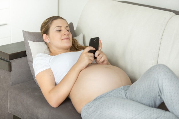 Beautiful pregnant woman lying on sofa and using smartphone