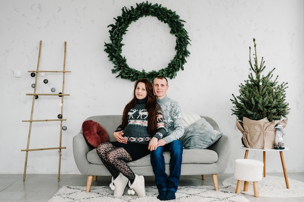 Photo beautiful pregnant woman and man in soft sweaters on sofa near tree