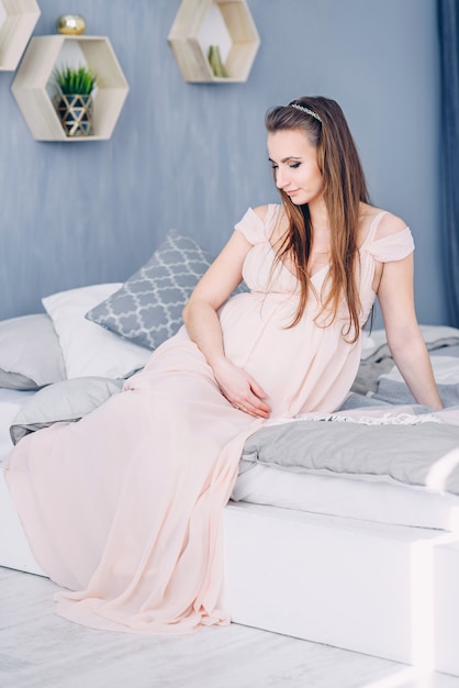 Beautiful pregnant woman in a pink dress sitting on the bed and holding pregnant belly