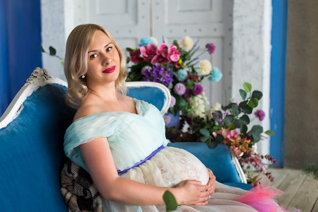 Foto una bella donna incinta siede tra i fiori in uno studio fotografico su sfondo blu in stile loft