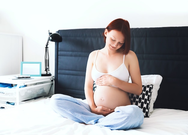 Красивая беременная женщина сидит на кровати и держит руки на животе в спальне дома
