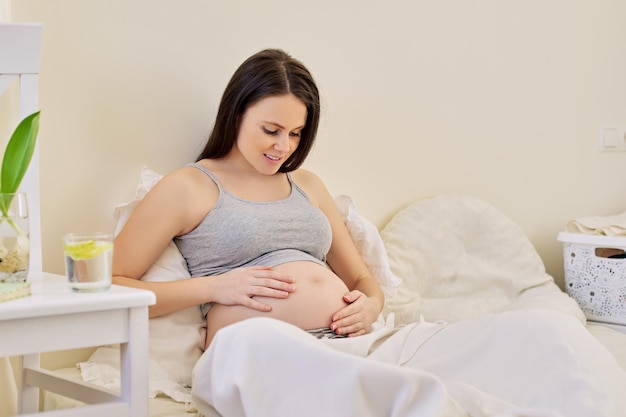 Beautiful pregnant woman sitting in bed at home touching belly, beginning of third trimester of pregnancy