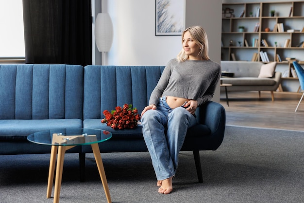 Beautiful pregnant woman sitting on couch in living room near beautiful bouquet