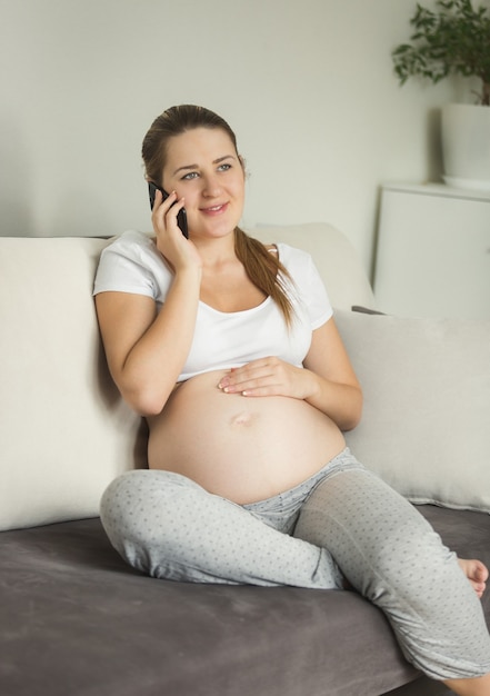 Красивая беременная женщина сидит на диване и разговаривает по телефону
