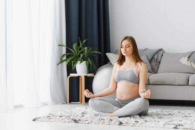 Bella donna incinta seduta sul tappetino da yoga in soggiorno, praticando la meditazione, giovane donna in attesa che fa sport a casa, godendo di uno stile di vita sano durante il periodo della gravidanza, copia spazio