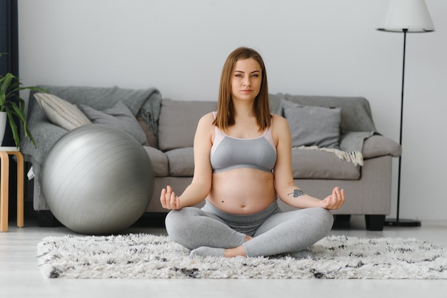 Beautiful pregnant woman sitting on yoga mat in living room, practicing meditation, young expectant lady doing sports at home, enjoying healthy lifestyle during pregnancy time, copy space