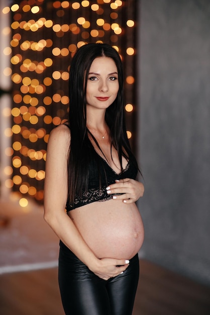 Beautiful pregnant woman stylish and happy pregnant woman Happy pregnant girl in a gray dress smiles joyfully in a dark studio on the background of New Year's lights