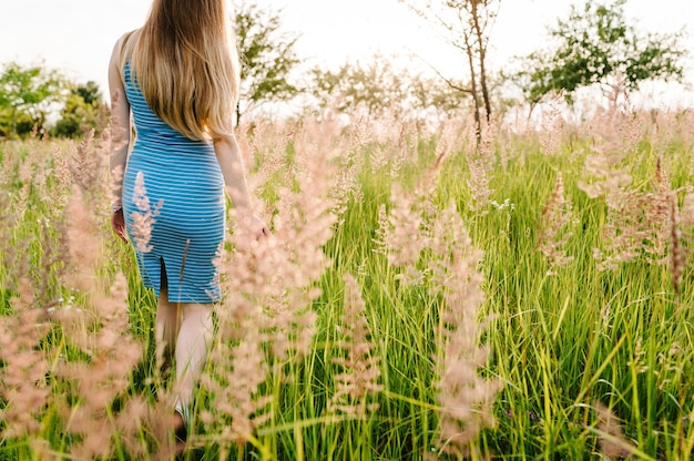 Bella donna incinta in abito estivo alla moda andare a piedi in campo con i fiori alla luce del sole, godendo la sensazione.