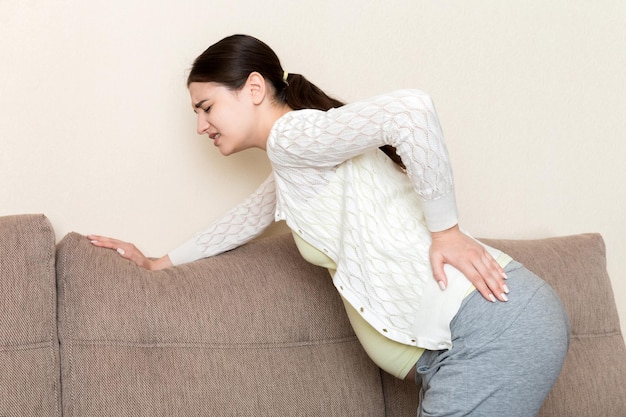 Beautiful pregnant woman touching her tummy and keeping one hand on her back at home on sofa